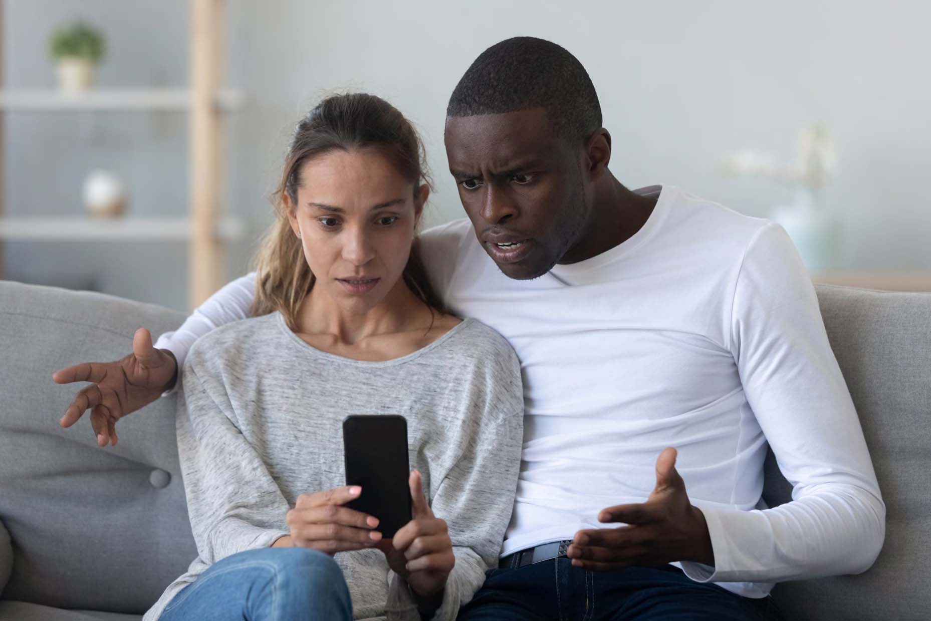 A man and woman sit on a couch, looking at a phone with a shocked expression.